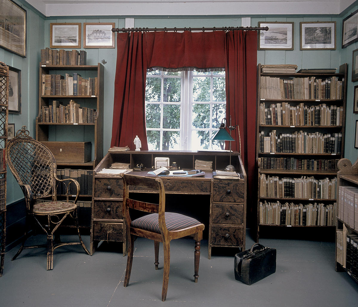 Interiør; bibliotek med bokhyller på begge sider av et vindu med røde gardiner, skrivebord og stol foran vinduet.
