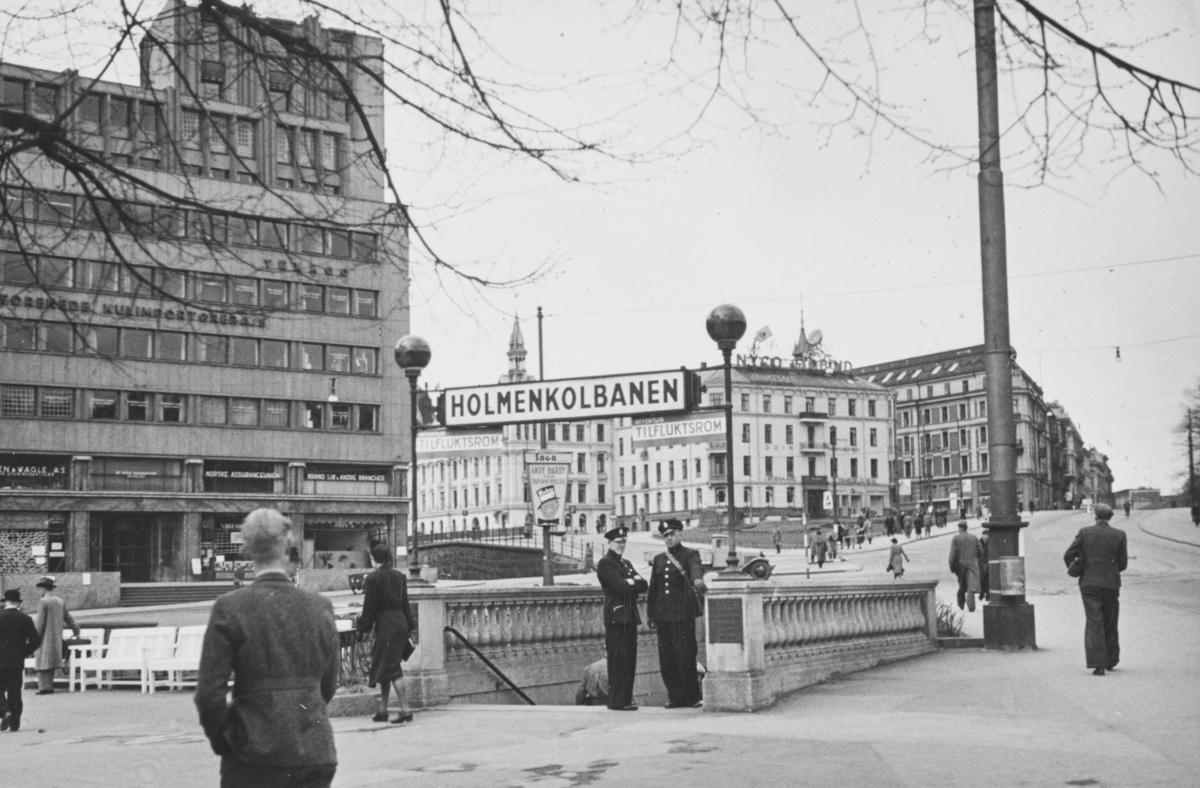 Ved nedgangen til Nationaltheatret stasjon i Oslo. Undergrunnsbanen ble benyttet som ffentlig tilfluktsrom under okkupasjonen. Politikonstabler utstyrt med gassmaske står klar. I bakgrunnen Victoria Terrasse, som ble bombet og delvis ødelagt 25. september 1942 og 31. desember 1944.
