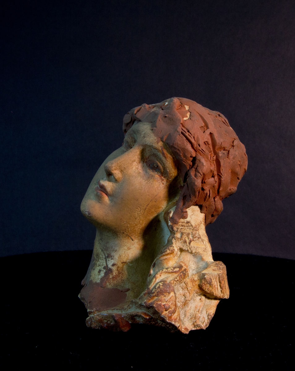 Studiemodell av gips föreställande kvinnohuvud. Kompletterad med plastelina av Julius Kronberg i form av hår och blomsterkransliknande del. Sekundärt färglagd och håret förstärkt med blyertsstreck.