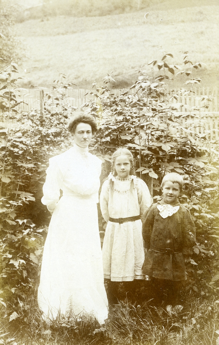 Prospektkort av to barn og ei kvinne som står ute i ein hage.