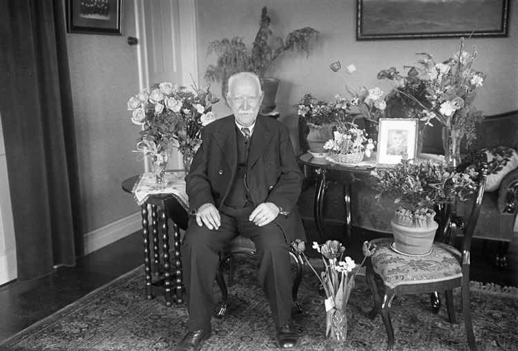En man sitter med blommor och presenter omkring sig efter en uppvaktning.I bakgrunden syns en soffa m.m.
