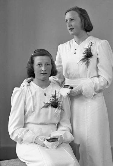 Foto av två systrar i vita konfirmationsklänningar. 
I händerna håller de små psalmböcker.
Ateljéfoto.