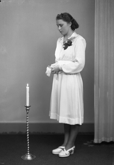 Foto av en ung kvinna i vit konfirmationsklänning. Hon står bredvid en tänd golvljusstake.
Helfigur. Ateljéfoto.