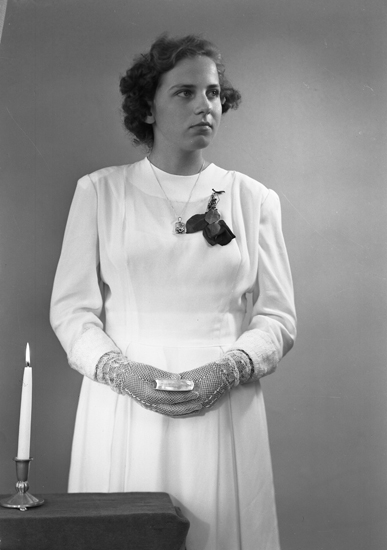 Foto av en ung kvinna i vit konfirmationsklänning och genombrutna vantar. I händerna håller hon en liten psalmbok.
Knäbild. Ateljéfoto.
