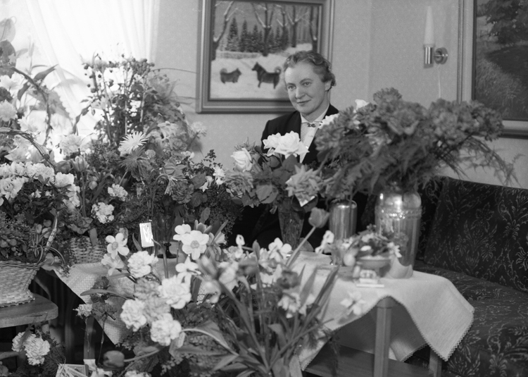 Foto av en medelålders kvinna, klädd i dräkt, som står i ett vardagsrum, omgiven av blommor.
Trol. fru Anna-Lisa Palmgren, Lekarydsvägen, Alvesta.
Källa: Telefonkatalog, Alvesta 1953.