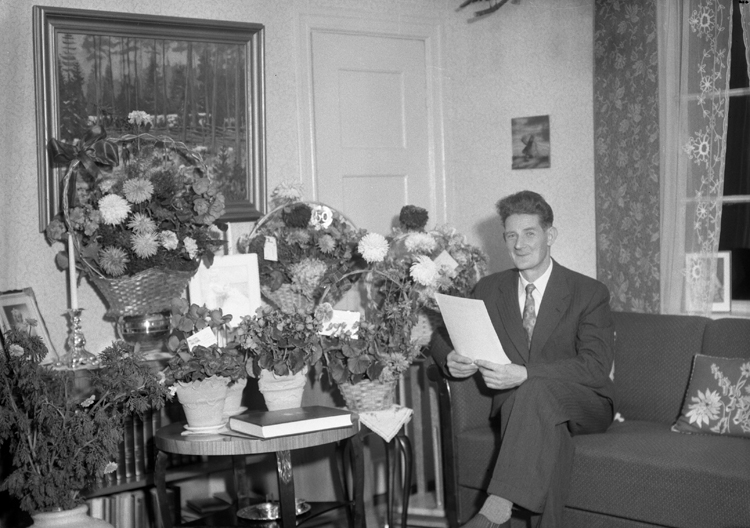 Foto av en man i kostym som sitter i ett vardagsrum med mängder av blommor bredvid sig.
Sven Einar Yngve Karlsson (23 oktober 1906-1982), Hjortsberga (senare Alvesta). Gift 28 februari 1931 med Teolinda Antonia, f. Lindahl (1908-1987).
Källa: Sveriges Dödbok 1901-2009.
