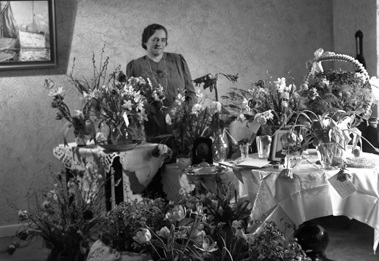 Foto av en medelålders kvinna i klänning som står vid ett bord fyllt med blommor, i ett vardagsrum.
Fru Elsa Wieslander, f. Davidsson (1 april 1894-1973), Alvesta. Gift 19 april 1924 med banktjänsteman Karl Uno Wieslander (1889-1958).
Källa: Sveriges Dödbok 1901-2009.