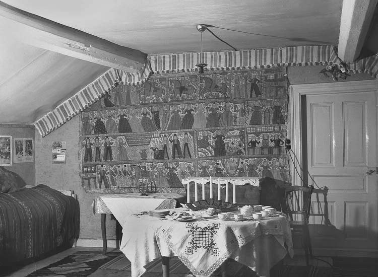 Interiör, med ett julbord och till vänster syns en soffa. På väggen hänger en 
stor bonad med olika bilder.