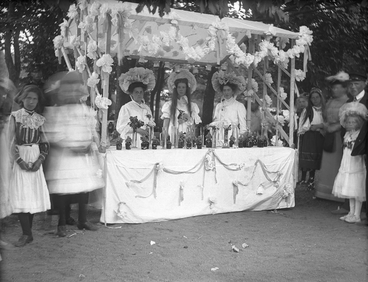 Foto av ett marknadsstånd, dekorerat med girlander m.m. Bakom står tre unga damer i ryschiga stora hattar. Framför dem står en mängd vaser med blommor.