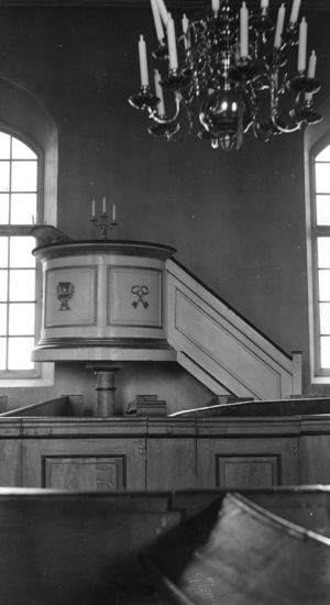 Foto på en predikstol med kyrkbänkar framför och en ljuskrona i taket.