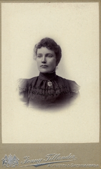 Porträtt (bröstbild), ateljé, av en kvinna i mörk, höghalsad klänning med en brosch/medaljong i halsgropen.