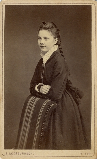 Porträtt (knäbild, halvprofil) av en ung kvinna, klädd i mörk klänning med stor pipad krage i vitt. 
Vid kragen syns en brosch med en smal klockkedja. 
Hon lutar armbågarna mot ryggstödet på en klädd stol.