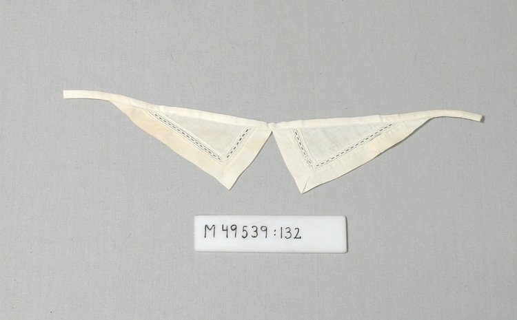 Stärkt vit krage av troligen linne, ev. bomull.
2 trekantiga "snibbar" som är fastsydda på ett band.
"Snibbarna" har en 10 mm bred infälld spets.
Bandet är avklippt i ytterkanterna, ofållade.


Inskrivet i huvudbok 2008-07-21