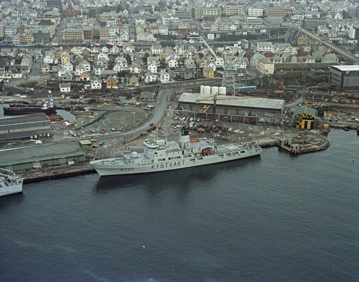 Flyfoto H.M.V. Kystvaktskipet W322 ligger til kai. Verkstedområdet og bebyggelse i bakgrunnen.