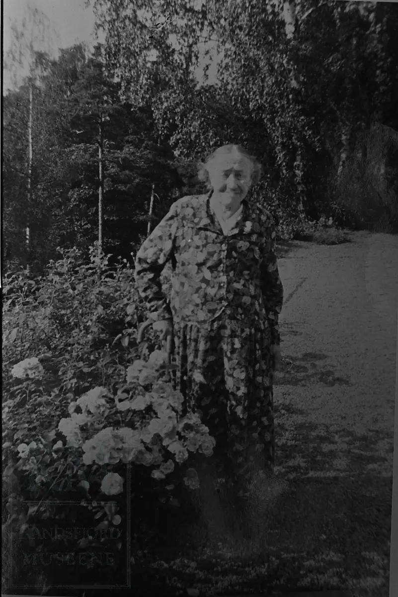 Eldre dame i blomstret kjole ved siden av et blomsterbed. Karen O. Foss