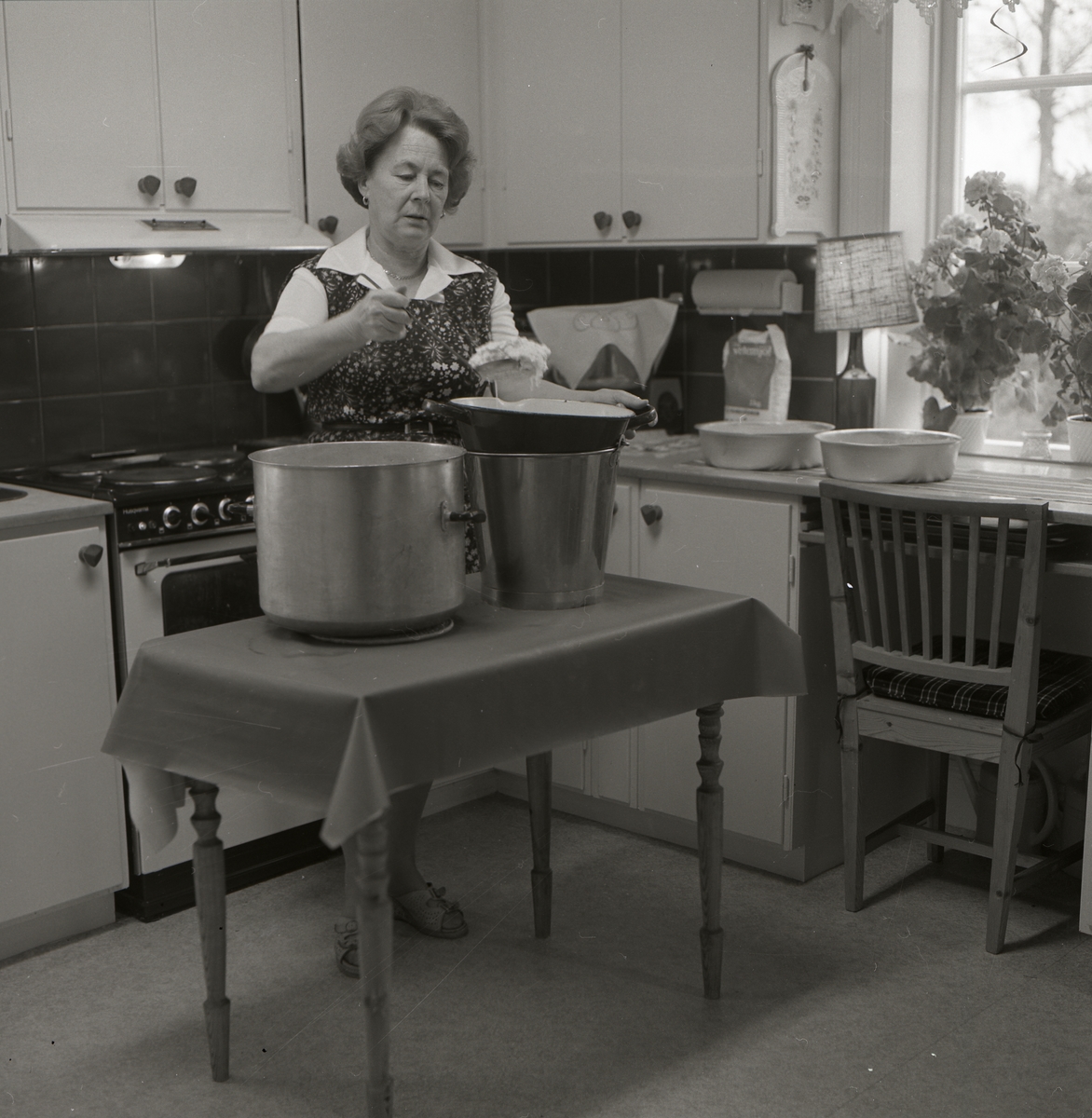 En kvinna står vid ett bord i kökets mitt och slevar ostmassan från en hink till en kastrull för att laga ostkaka. Bakom henne skymtar kökets spis fram och till höger ligger två formar redo för att fyllas och gräddas.