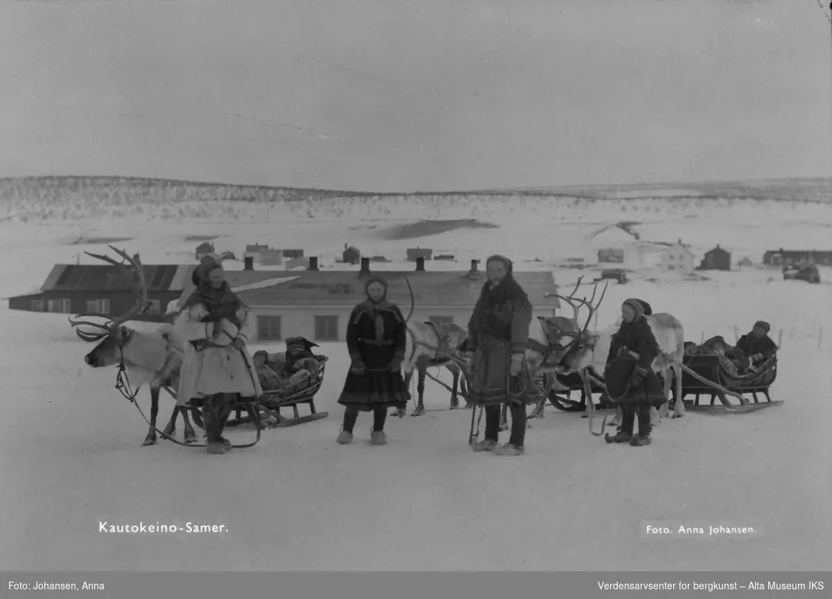 To samiske kvinner og fire barn fotografert utendørs på vinteren sammen med reinsdyr. To sleder det sitter barn i. I bakgrunnen ser vi bebyggelse.