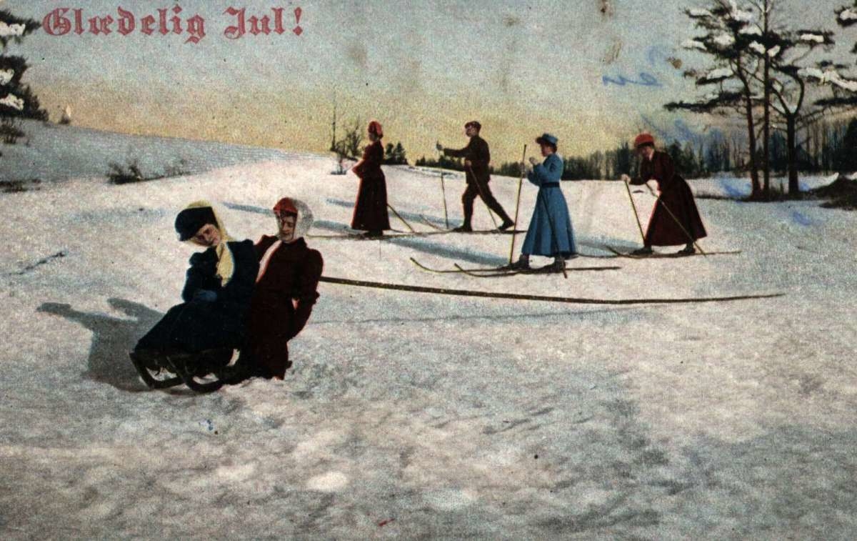 Julekort. Nyttårshilsen. Kolorert fotografi. Vintermotiv. Landskap med skiløpere i bakgrunnen. Kvinner på kjelke i forgrunnen. Stemplet 1910.