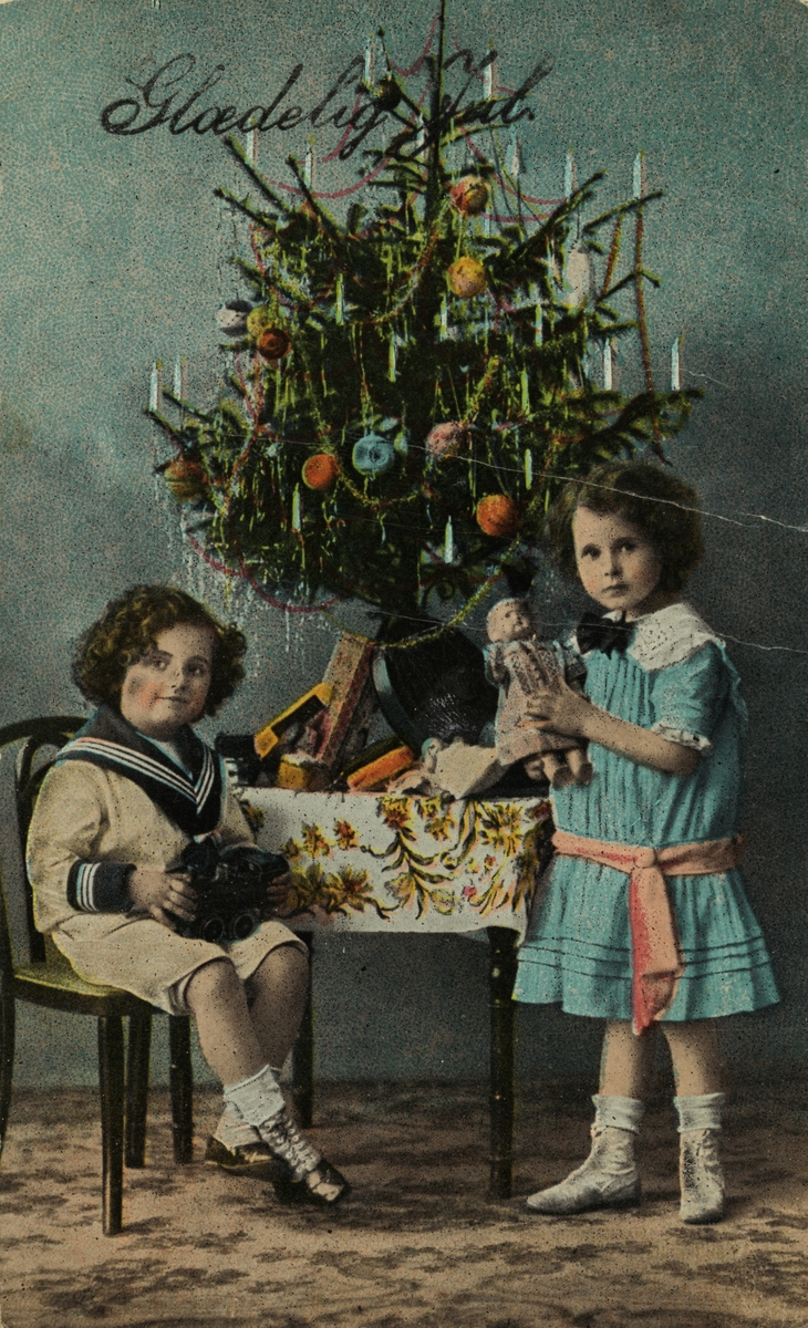 Julekort. Jule-og nyttårshilsen. Pike og gutt med gaver under juletreet. Gulldekor. Håndkolorert fotografi. Utenlandsk kort.