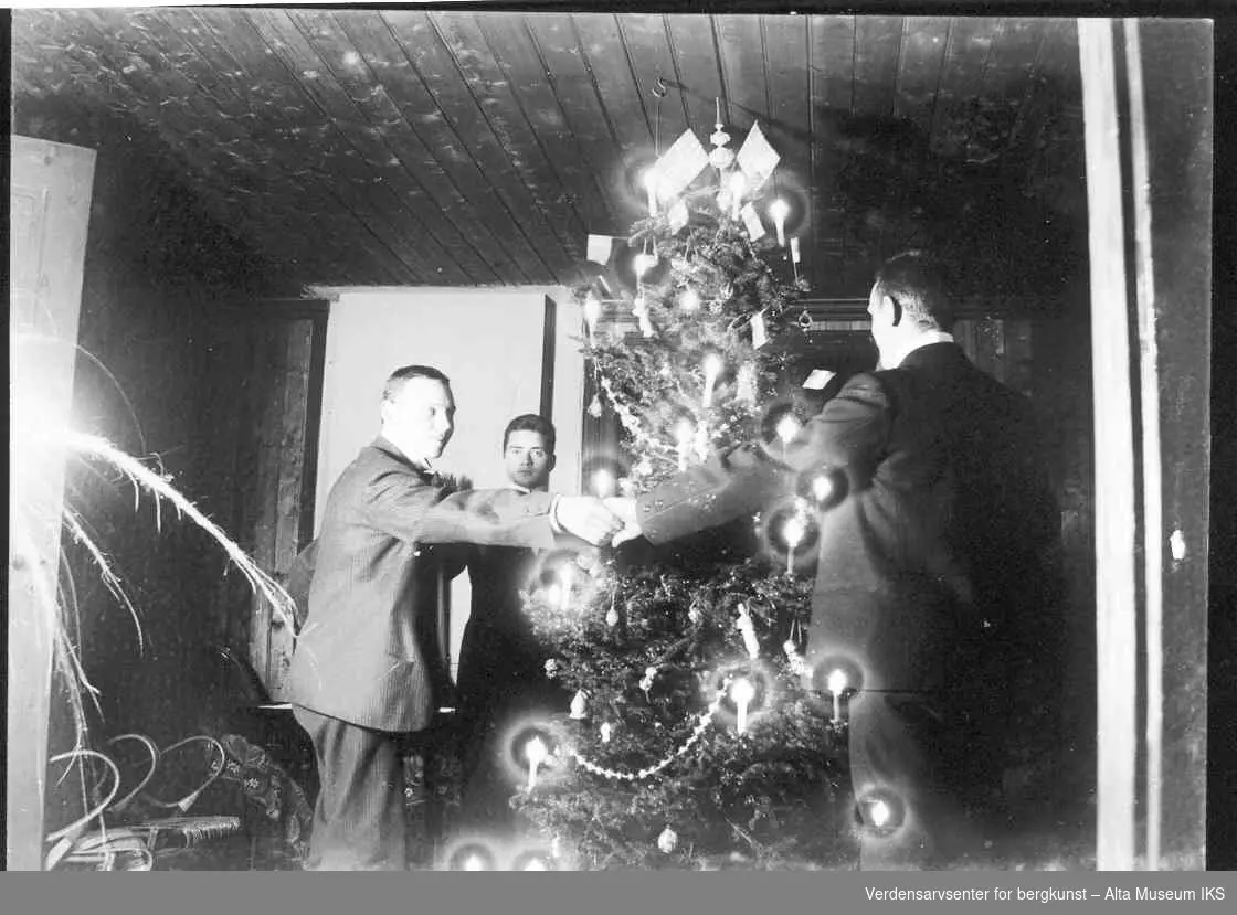 Tre menn går rundt et pyntet juletre i en stue.