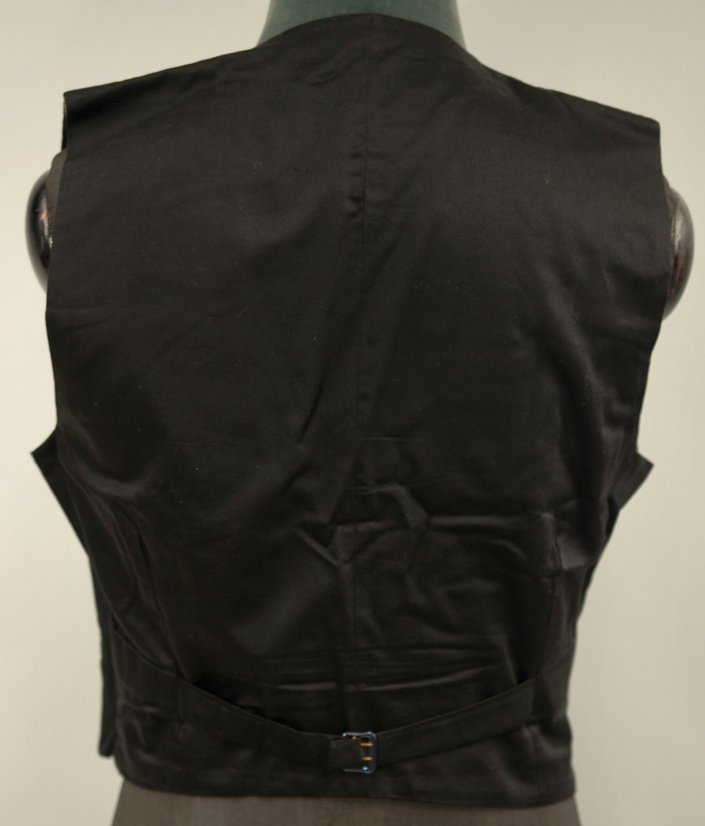 A: Jakke, Svart klede, krage med silkeslag. Enkeltspent, 1 trekt knapp, 3 trekte knapper på kvar arm. Svart silkefor. L.71,5 cm bredde ved skuldrane 45 cm
B: Bukse, svart klede, silkeripsbånd i sidene. L. 103 cm, Vidde nede 24 cm
C: Vest, svart klede, svart silkefor i rygg, foret med lystripet sateng. Enkeltspent med 4 trekte knapper, 4 lommer. L.48,5 cm x 38 cm
D: Sløyfe, Sort silkerips 55cm x 3,5 cm