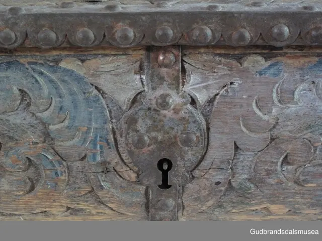 "Bladformer, smøygde band og drageornamentikk. Millom kantjarn og utskurden hev lokket fint utskorne små blad og halvrunde aggar. Vel vedlikehalde, sterk jarning og prydelege gangjarn. Truleg frå 1500 talet. Fra Haugen, Øystre Gausdal". (Jf. "Katalog for Lom Bygdesamling,1925).
Skrinrammen er holdt sammen av utvendige hjørnekantbeslag; jernhandtak i hver side. Fronten har nøkkelhullslås med beslag rundt. Lokket er satt samen av 2 bord, forsterket med jernbeslag rundt kanter på overside; to hengselbeslag festet til innside av lokk og bakstykke. Skrinsider blir holdt sammen av beslag ned langs hjørnekanter; har jernhandtak festet på jernbeslag.
Bunnen er satt sammen av 2 bord, festet til hverandre med to forsenka treplugger; bunnen var opprinnelig festet til underkant av skrinsider med 4 x 4 trenagler,seinere festet med spiker. 
Det er jernbeslag  på bakstykket midt, rundt hjørnekanter, omkring nøkkelhullslås, rundt lokk samt 2 handtak med beslag. Alle beslag er festet med store jernnagler; beslag på sidestykker er festet med små jernnagler under skrinbunnen. 3 av 8 bunnfester for beslag er fortsatt intakte.
DEKOR. De store jernnaglene med forskjøvet plassering på beslag ble sannsynligvis brukt som dekorelement; runde beslag på sider, bakstykke og front. Flatskurd på lokk med flettet, stilisert renessanseranke, der bladverk er "omgjort" til småspiraler eller rundinger.  På skrinsidene akantusskurd med c-former, bladtunger uten stilk og naturalistiske småblad i rokokkostil. Flatskurd var vanlig i Gudbrandsdalen i perioden ca. 1650-1750, mens akantusmotivet først ble vanlig på bruksgjenstander i løpet av siste halvdel av 1700-tallet. Den mørkere grønne og blå dekorfargen var vanlig i siste del på 1700-tallet; den brune bunnfargen var vanlig i bruk fra 1780-tallet. 
BRUK. Før kofferten kom i bruk hadde man reiseskrinet, som ble brukt til oppbevaring av bl.a. klær, verdisaker og mat på reiser. Skrinet hadde ofte et eller to mindre, smale rom langs den ene skrinsiden og ett større rom. De fleste reiseskrin var i tillegg forsterket med jernbeslag langs alle kanter og med en solid nøkkelhullås til sikring mot støt, slag og tyveri.