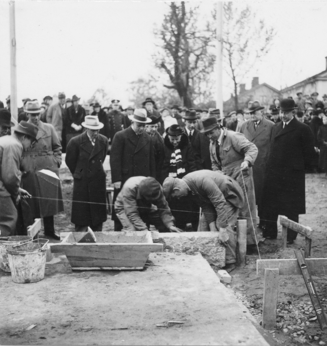 Grundstenen för Gävle Museum lägges 26 oktober 1938. Några byggnadsarbetare är i färd med att slutföra arbetet för dagen. Borgmästare Nils Berlin m.fl. övervakar arbetet.