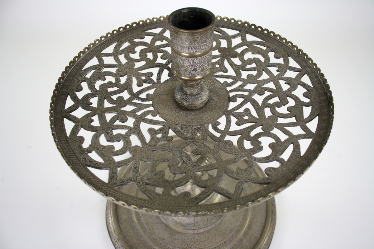 Moskélampa av mässing med rik dekor och stor rund fot. Ljushållare och under den rund platta med genombrutet mönster och dekor bestående av citat ur koranen på arabiska. Persien.