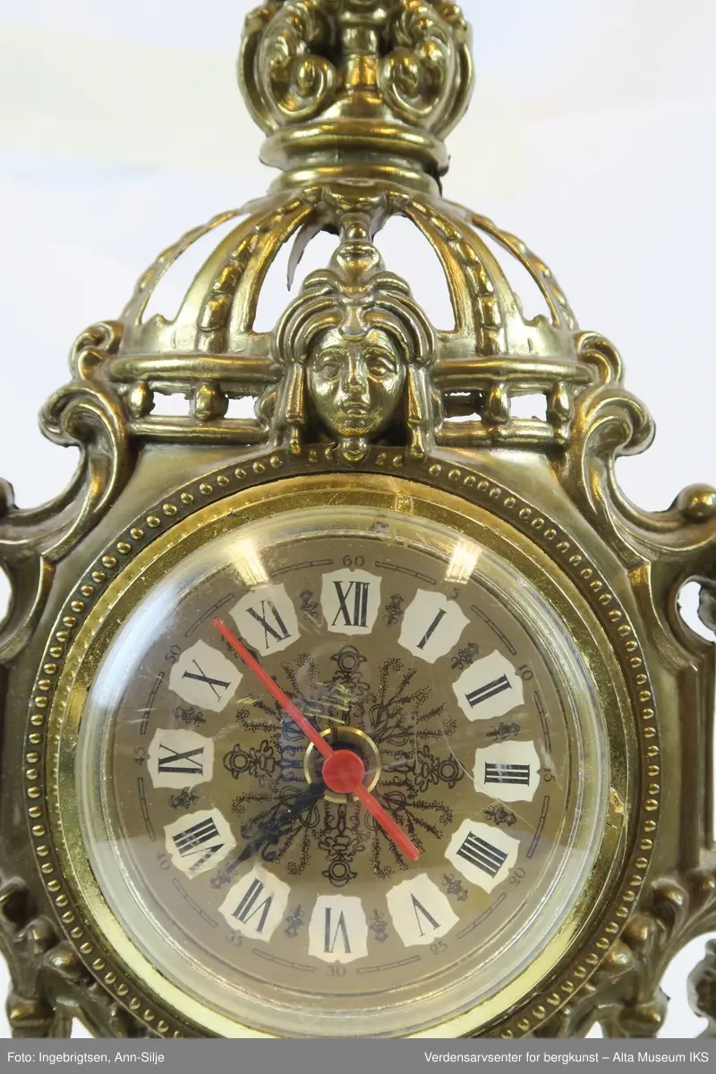 Batteridrevet ur. Stående oppsats med barokk innfatning og innfelt urskive med romertall.
