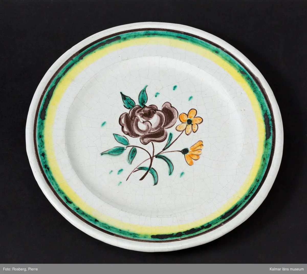 KLM 45844:2 Fat, av keramik. Runt, med blomma i lila, rosa och grönt, grön rand på brättet. Signerad, J.S. Målad och signerad av John Sjöstrand, tillverkad vid Sandbäcks kakelfabrik AB. Dateras till slutet på 1920-talet till 1931.