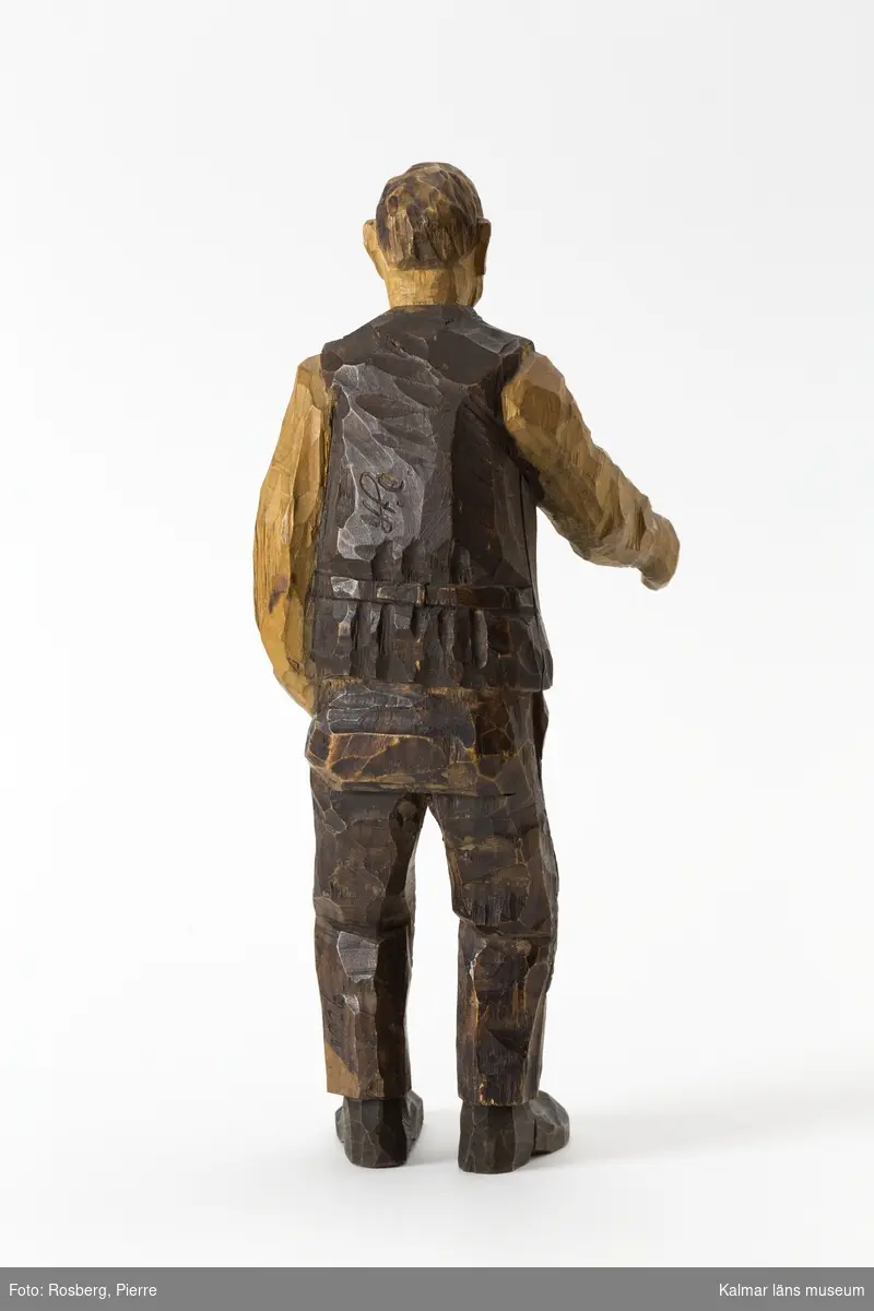 KLM 45932:1 Skulptur, av trä, målad. Figur i form av en stående man. Han har vänster hand i byxfickan och den högra armen sträcker han fram, möjligen har han ursprungligen haft något föremål i den framsträckta handen, barhuvad, klädd i skjorta väst och byxor, stora lappar på knäna, och skor. Signerad på ryggen, H.C., på vänster byxben inskuret, 1926. Målad i bruna nyanser. Tillverkad av Helge Rugland, f. Carlsson.
