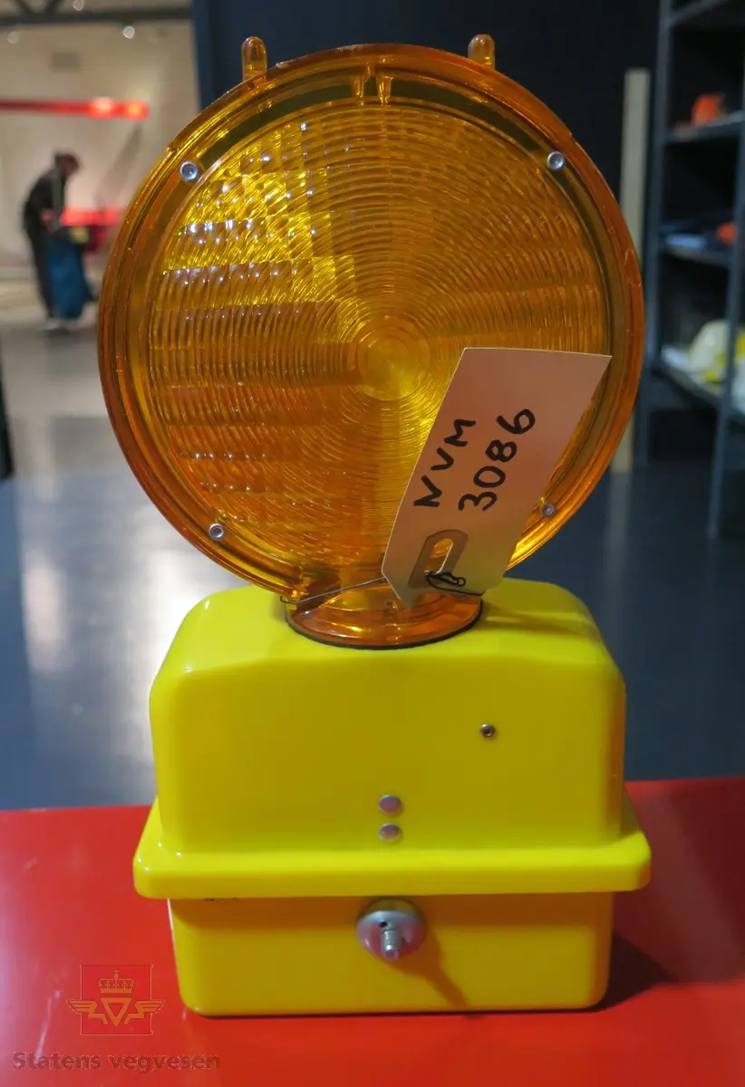 Varsellampe, blinkende type, blinker i to retninger. Lagd av gul plast, med oransje plastkuppel på toppen. Drives av 6 volt batterier, disse mangler. Lampen startes og stoppes ved å stikke en ståltråd eller lignende gjennom et hull i lampehuset.