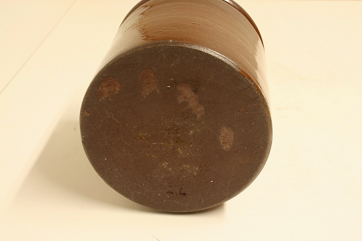 Sylinderformet sylterkukke med profilert munningsrand. Den er laget av brunglasert keramikk av rød leire.
