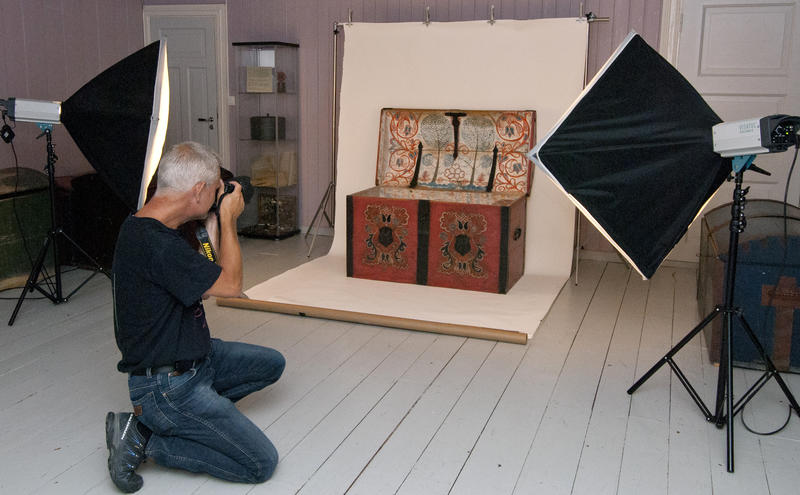 Gammel kiste er plassert på hvit fotobakgrunn, blitslamper står vinklet mot objektet, og en fotograf står på kne med håndholdt kamera og tar bilder av kista som er den del av museumssamlingen.