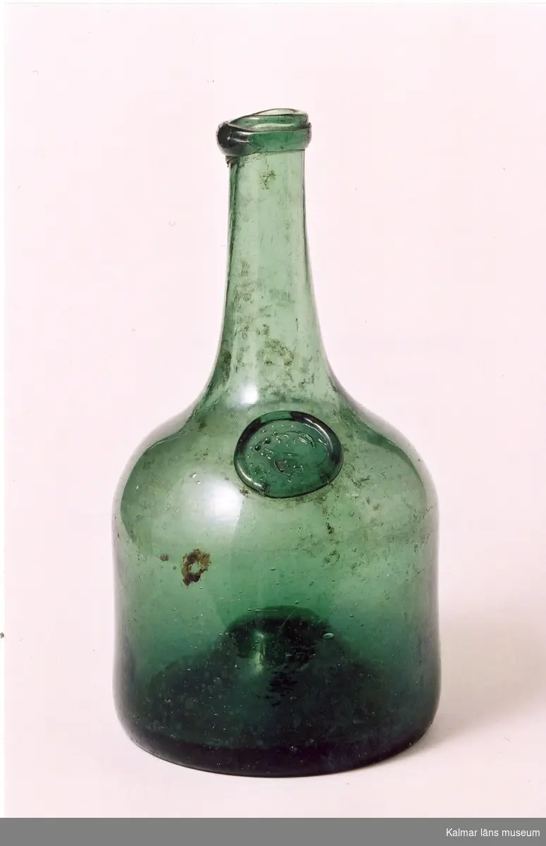 KLM 24961 Flaska, av grönt glas. Balongflaska. Hög kulle i botten. På skuldran ett glassigill. På sigillet stämpel: 1/3, L W G B. (LångWiks GlasBruk). Datering, omkring 1800-1815(?).