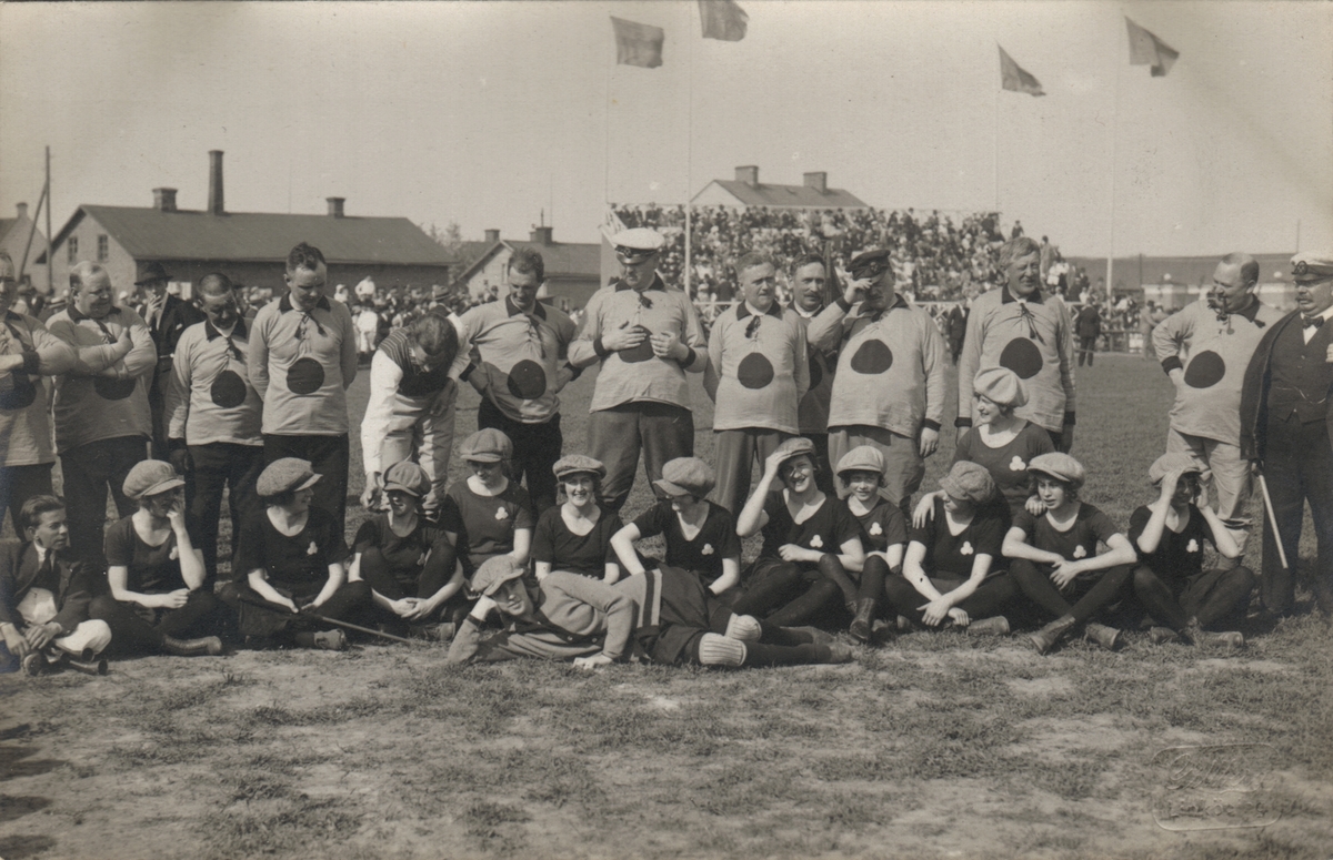 Bilden visar en laguppställning till en fotbollsmatch under Barnens Dag 1922, Linnévallen. Stående är det Old Boys och sittande Kvinnliga Gymnasikföreningen. Kortet är taget på en gräsplan med publiken på läktaren i bakgrunden tillsammans med de vajande flaggorna.