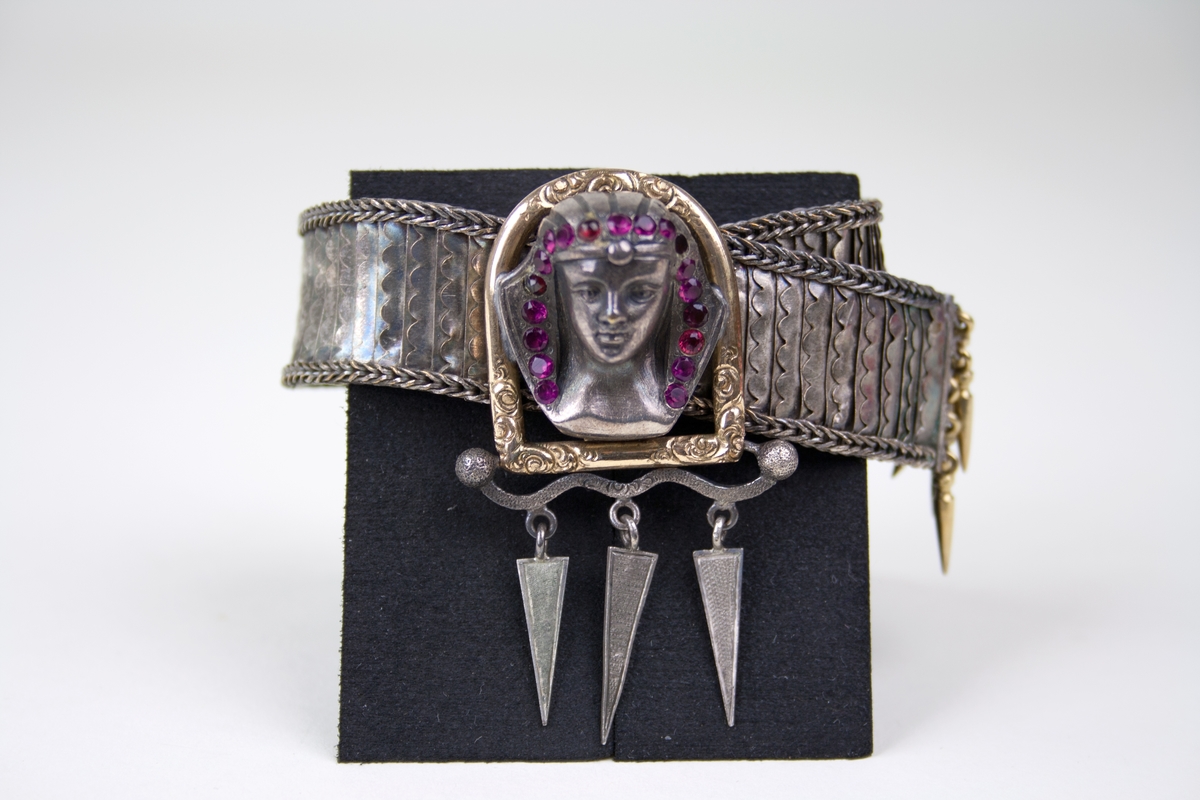 Armband av silver med spänne i form av ett egyptiskt kvinnohuvud av guld, silver och rubiner. Bandet är format av små lodrät ställda silverfjäll och flätat kantband. Under spännet och på bandänden hänger triangulära berlocker. Armbandet saknar stämplar.