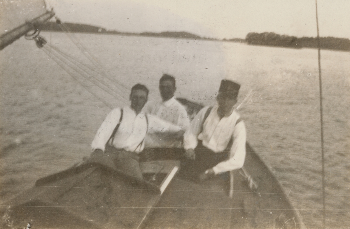Tre män i segelbåt. I bakgrunden syns en holme och strandlinje.