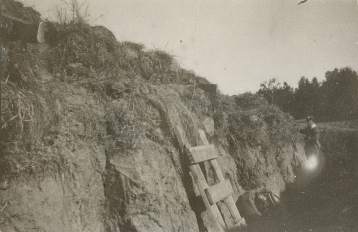 Skyttegrav inifrån med nedgrävt dike och vallanläggning samt en stege som leder till vallen. I bakgrunden står en soldat.