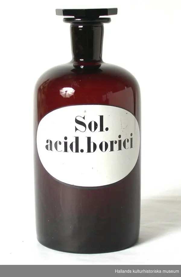 Apoteksflaska av glas. Brun. Cylindrisk form. Med slipad propp och kort smal hals.a) Flaska. Höjd 24,5 cm, diameter 10,5 cm (i bottnen), diameter 2,5 cm (i halsen). Med signatur: "Sol. acid. borici". Märkt i bottnen: "1075". b) Propp med åttkantig hätta. Märkt: "15".