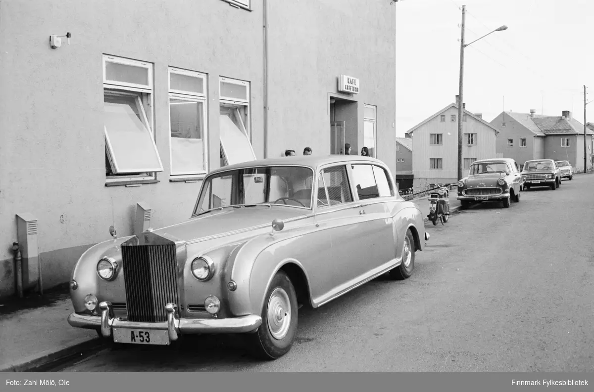 Bildeserie med biler fotografert av Ole Zahl Mölö i perioden 1967-69. 

1967: En Rolls Royce (med registerskilt A-53) er  parkert utenfor en lokal kafé i Vadsø. Bilen var en av to stykker eid av den britiske ambassaden. Ambassadebilen var i Vadsø i anledning en rundreise som den britiske ambassaden foretok dette året. Ambassadørene skiftet ofte i 60-årene og de skulle bli kjent med landet. Sjåføren som kjørte den staselige bilen het Ronald Mansell. Han hadde kjørt bilen helt fra Oslo til Vadsø. Ambassadørene derimot, tok fly og ble kjørt rundt i nærområdene når de landet. Ronald Mansell hadde blitt anbefalt spisestedet og har senere fortalt at maten var både billig og god. Ronald Mansell ble godt kjent i Norge på disse turene.
Ronald Mansell ble  født i Sheffield 16.04.1921. Han vervet seg i RAF 2.04.1941 og ble bakkemannskap for fly / flyplass. Han var blant de første som kom til Norge i 1945 etter å ha vært stasjonert på Island. Soldatene var stasjonert der uten å vite at de skulle videre til Norge. Mansell var da i en spesiell avdeling for transport. Han traff sin norske kone i 1946 dro til England 2.08.1946 for å dimittere. Da han kom tilbake og giftet seg med henne.  De fikk en sønn 01.02.1948 og i mars, samme år,  fikk han jobb som privat sjåfør for ambassadøren.  Ronald Mansell og familien bodde på den britiske ambassaden i Oslo frem til 1980.
Mansell ble tildelt British Empire Medal for sitt arbeid. På rekke og rad etter Rolls Royce-en står det parkert en Opel Rekord Caravan stasjonsvogn fra1958-60 (med registerskilt Y-22340),  en motorsykkel og en Ford 17M. Man kan skimte mennesker som står utenfor kafeen og vi kan se at vinduene er åpne i kafeen, så man kan regne med at en Rolls Royce vakte oppsikt i Vadsø. 