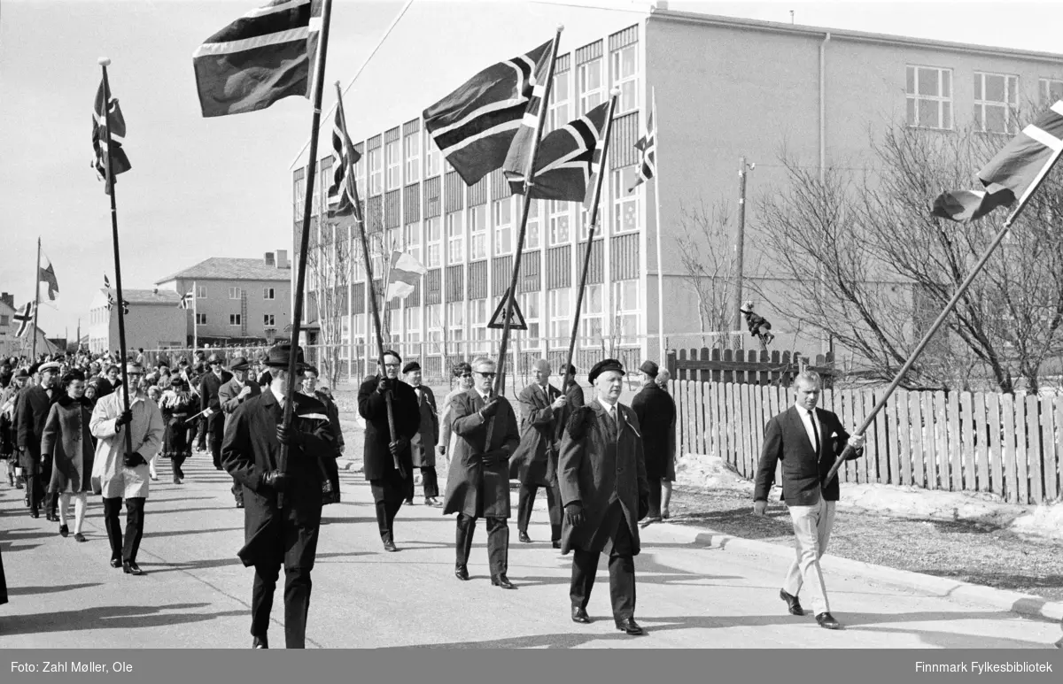 Vadsø 17.5.1969. . Fotoserie av Vadsø-fotografen Ole Zahl-Mölö. Hele 6 flaggbærere leder an i borgertoget. Etter flaggene å dømme er det god vind og skikkert en utfordring å bære flaggene. Litt lenger bak skimtes også et finsk flagg i toget.