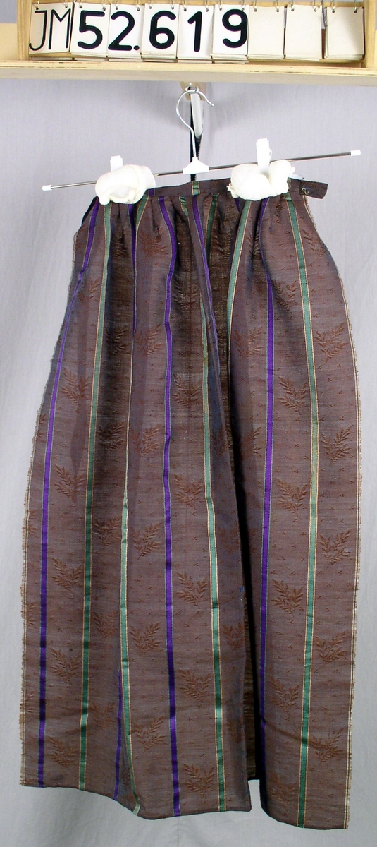 Förkläde i mönstervävt  brunt linnedamast med längstgående ränder i grönt och lila. Enligt påsydd textetikett: "Förklädet från Rydaholm 1850. Har burits till en brudklänning av damast - tyget finns på N. M. Julia Fredricsson".