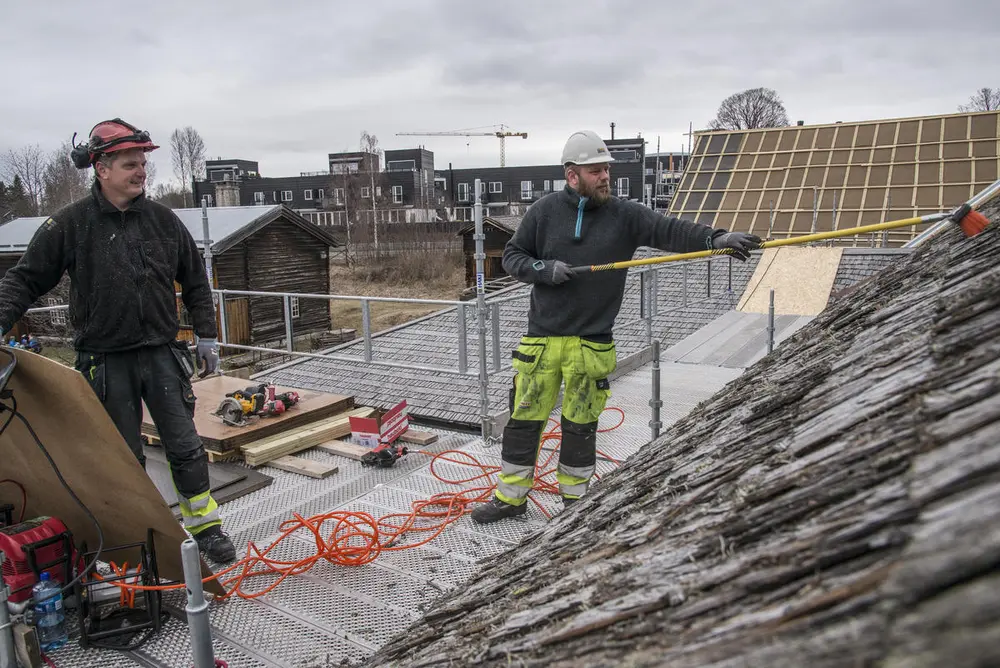 Menn med hjelm står på stillas og rengjør gammelt, flislagt låvetak for å legge på nye takplater.