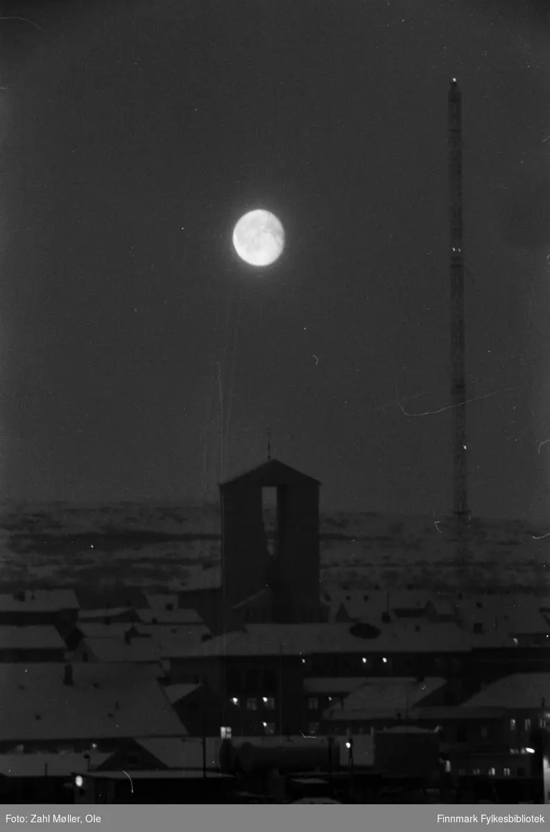Fotoserie fra Vadsø, april 1968. Fotografert av Vadsøfotografen Ole Zahl-Mölö. Månen lyser over Vadsø kirke og byen, fotografert om natten. Vi skimter radiomasta og lys fra bygninger i forkant av bildet.