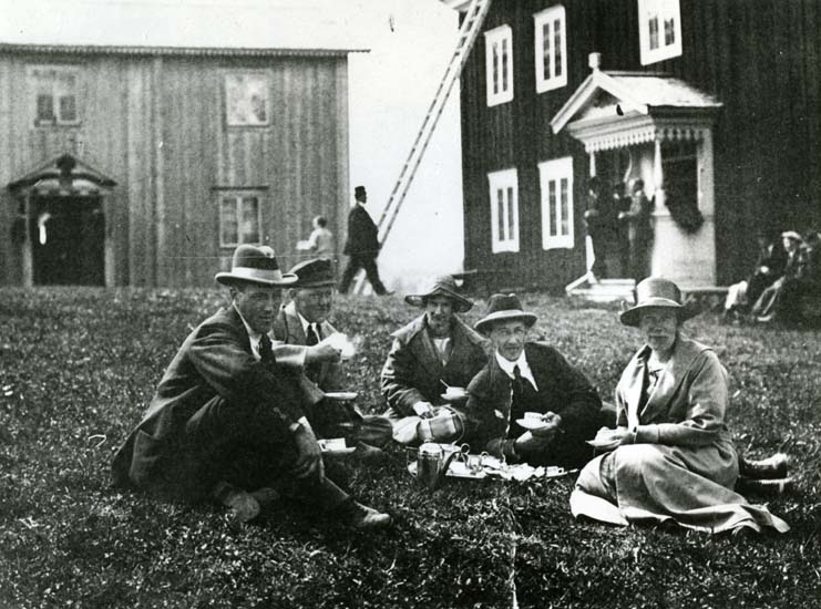 Vid Mårtensgården, Hembygdsgården i Edsbyn. En grupp människor sitter i gräset framför gården och dricker kaffe.