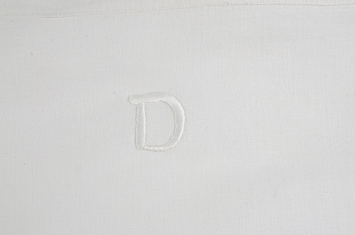 Underlakan av vit bomullslärft med monogram D i klumpsöm. Maskinfållat