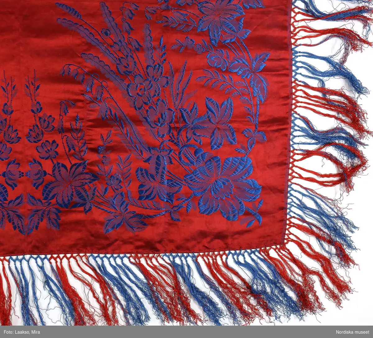 Kvadratiskt halskläde av sidensatin, röd botten med jacquardvävt blått blommönster, i hörnen buketter med långa stänglar med liljekonvaljer (?) och blåklockor, från buketten utgår slingor som möts på sidorna i smäckra blomstänglar , 2 maskinsydda fållar. Tvåknuten krusad  frans av tvinnat silke i omväxlande blått och rött. Mycket svag röd stämpel i ena hörnet med texten: Casparsson & Schmidt Stockholm.
Från Talma lappby. Vanligt bland samerna att ge silkekläden i friargåva. Denna  är en gåva från samen Johan Turi till konstnärinnan Emelie Demant Hatt som vistades och målade i sameland. Tavlor av henne finns i Nordiska museets samlingar.
I nyskick , troligen inte använd. Samma mönster finns från andra håll i landet. Casparsson & Schmidt var tillsammans med K.A.-Almgrens sidenväveri de enda svenska sidenväverierna i slutet av 1800-talet och deras dukar såldes över hela landet. C&S upphörde 1904.