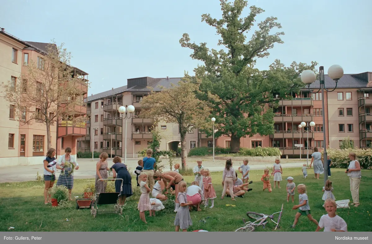 "”Stockholm Enskede gård”Barn och vuxna med cyklar och barnvagnar i gräset mellan hus. Stort träd i fonden."