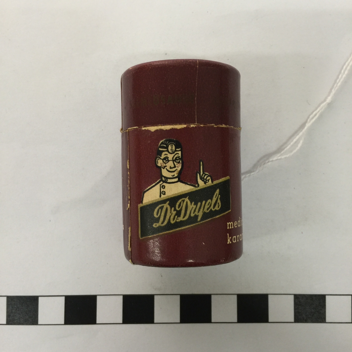 Cylinderformad tändsticksbehållare av papp i form av reklamartikel för Dr Dryels medicinska karameller.
Löst lock.